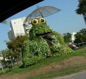 פסל חוצות באשדוד: צפרדע עם שמשיה - שלד רשת עם פרחי העונה. (צילום: רבקה יניב)